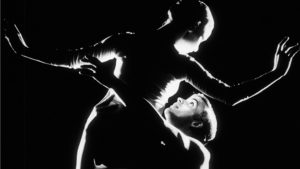 Black and white live-action film still. The outline of a woman being lifted by a man in a ballet pose is shown at close distance against a stark black background. The facial features of the man, on the right, are visible.
