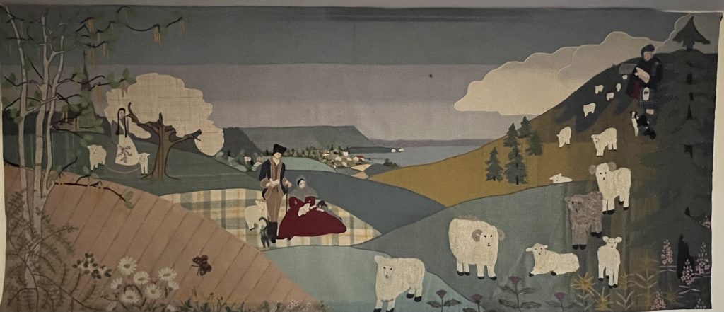 wool applique artwork with sheep grazing in a landscape and a couple in the middle ground. A woman stands under a tree with two sheep on the left hand side.