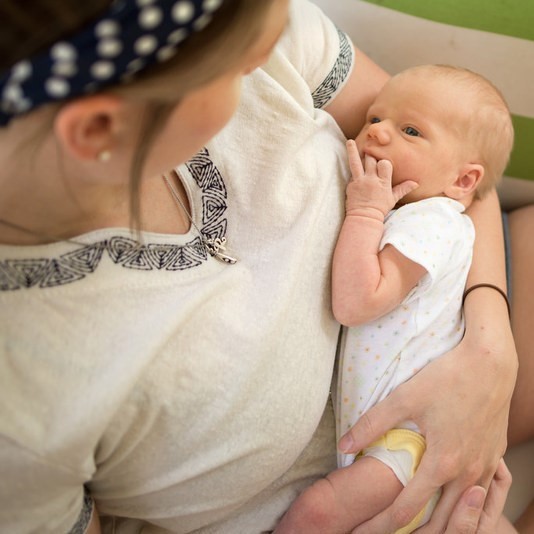 A young infant looks at a caregiver’s face while sucking it's finger. You cannot see the caregiver's face, as the caregiver is looking at the infant in return. They appear to be making eye contact with one another.