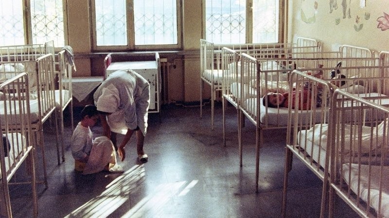 An image taken from an institute in Romania in 1992. A caregiver is bent over a child who is sitting on the floor. They are in a walkway between rows of cribs, some of which have children in them. There are large windows in the room, and what appear to be pictures of flowers on the walls that the cribs are up against.