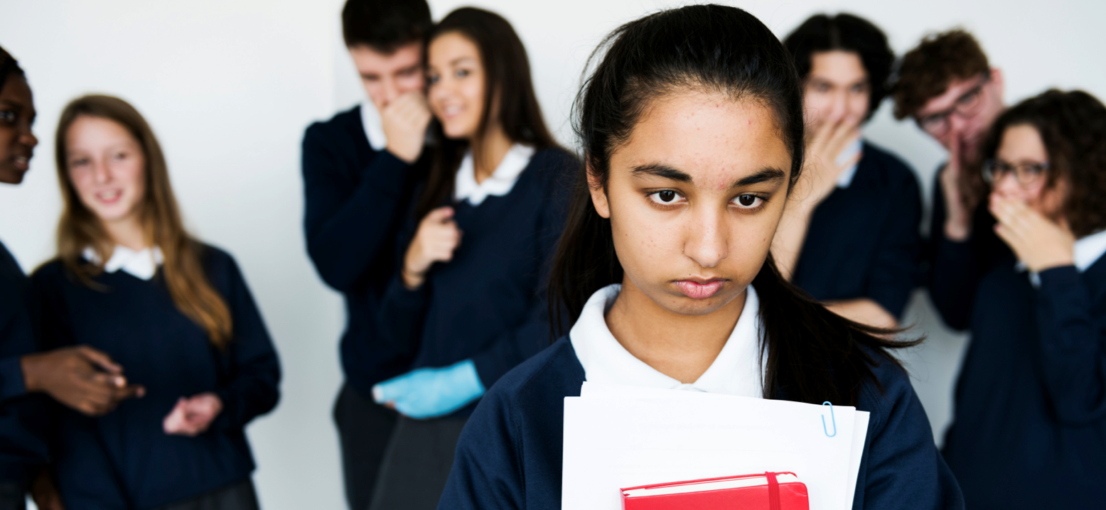 An image focused on the face of an adolescent girl with other adolescents whispering behind her and looking at her. The others in the background appear to be whispering about the girl.