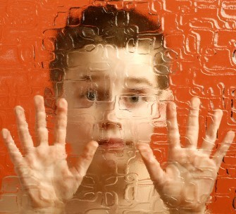 An image of a child in front of an orange background. The child has their hands up on a plastic wall. The wall is infront of the child, and is blurring the view of the child.