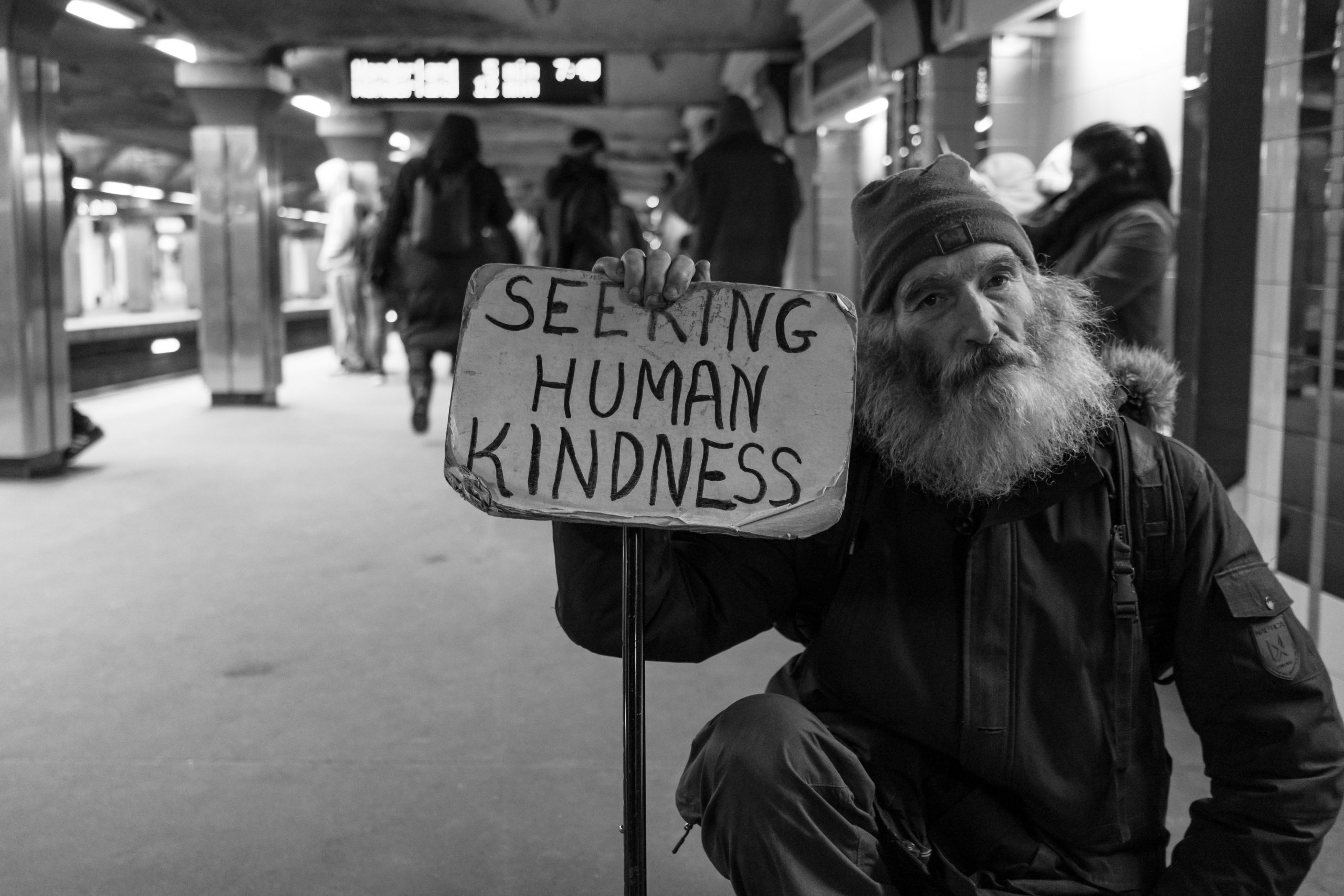 Elderly bearded man holding a sign, "seeking human kindness", in a subway station.