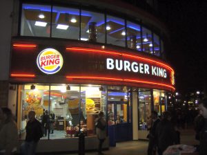 Exterior, at night, of a busy Burger King restaurant. The signage is neond with the BK logo and name prominently displayed.