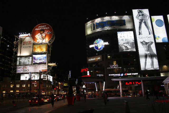 Night scene of a busy intersection with sides of skyscrapers all showing different sizes of advertising from a few feet tall to multiple stories. 