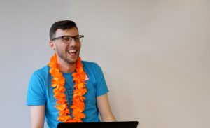 Adam smiles while standing at a podium and wearing a blue t-shirt and an orange lei. He is emceeing an event for children and youth.