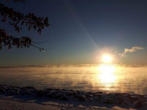 Photograph of a sunrise with blue sky over a lake in early winter. Snow covers the shore and steam rises off the water, obscuring the distant shore.