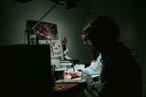 Low light photography of woman in gray knit sweatshirt writing on desk.
