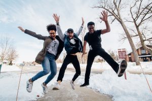 Three people jumping on ground near bare trees during daytime.