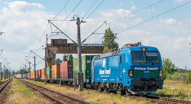 Blue train with shipping containers under a loading device in a railyard.