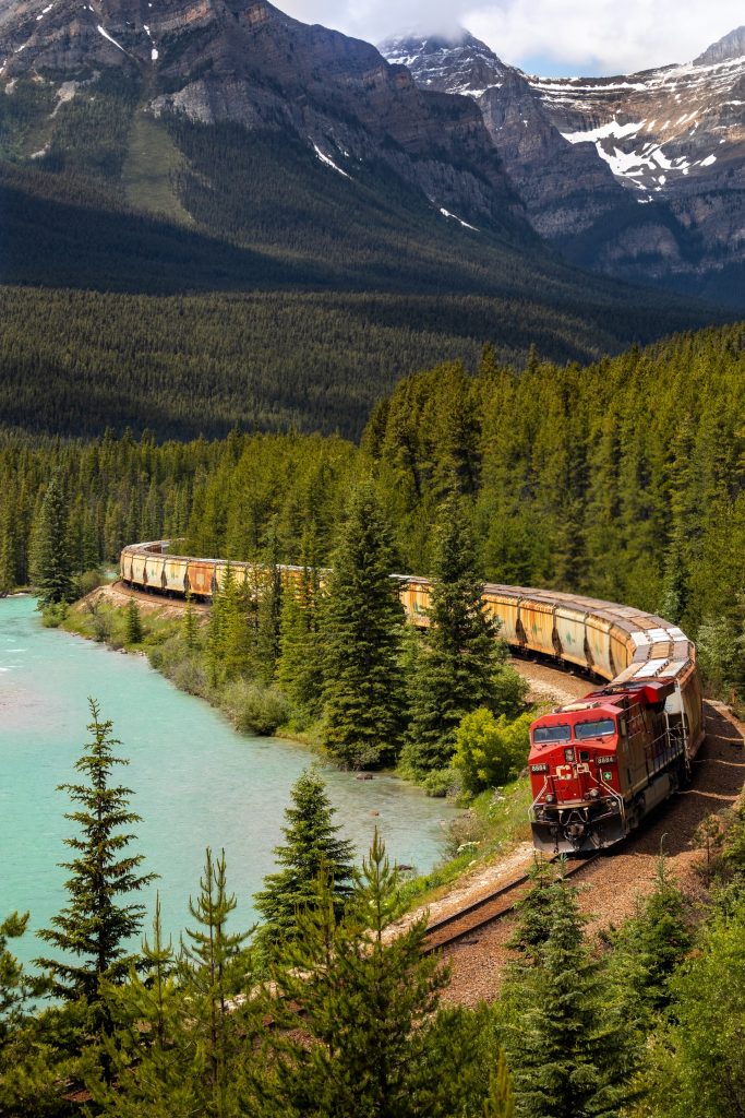 A red train pulls cargo past a body of water in the mountains on a sunny day.