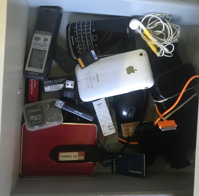 Open drawer with various obsolete electronics in it including a voice recorder, iPod, Blackberry, headphones, charging cable, and various memory cards.