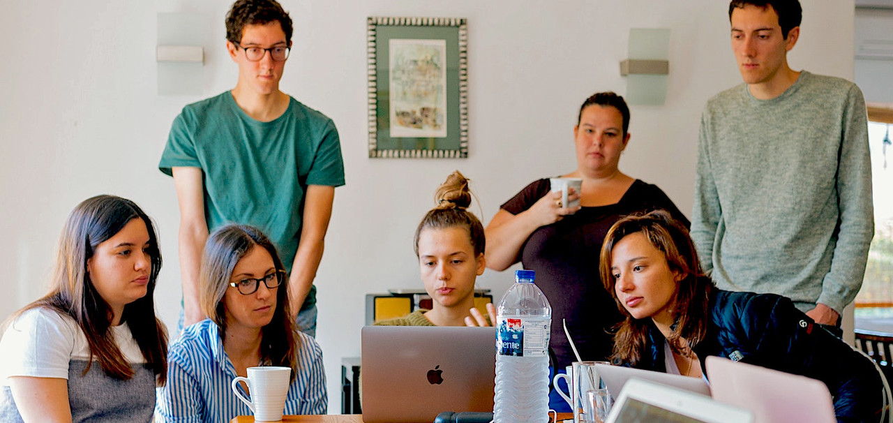 Image of 7 young adults gathered around a laptop in an office setting.