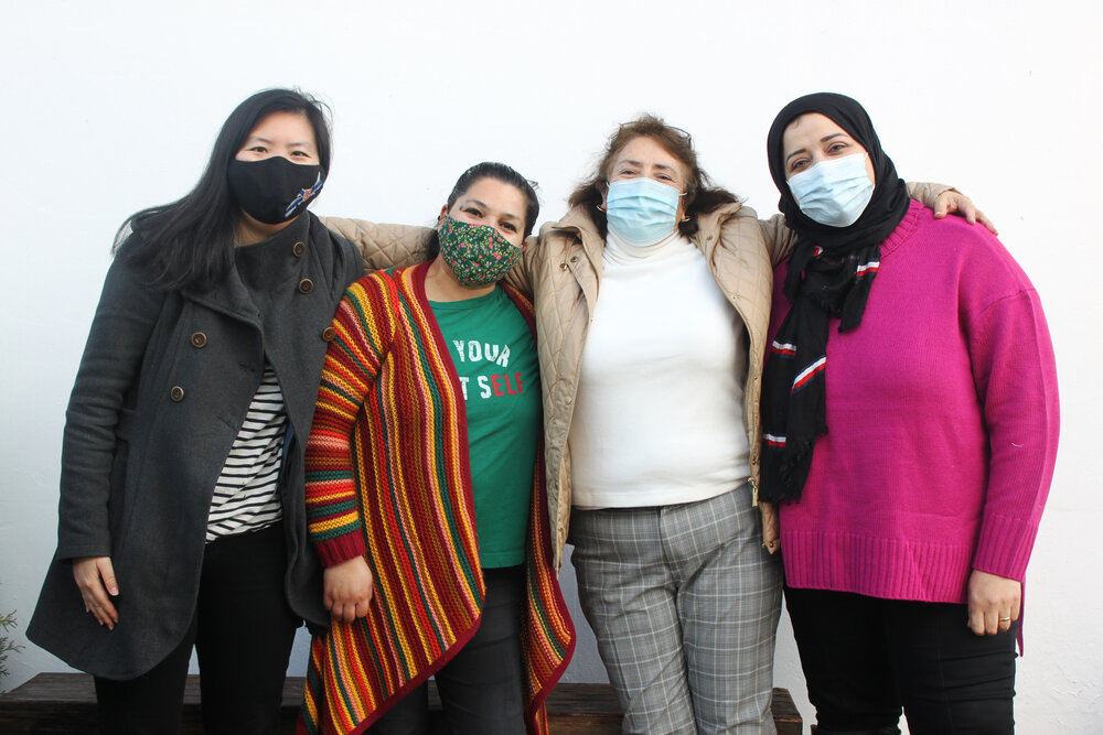Four women with arms on each others shoulders. Wearing COVID masks.
