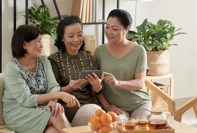 Positive elderly women gathered at home to drink tea, discussing news and show photos on smartphone