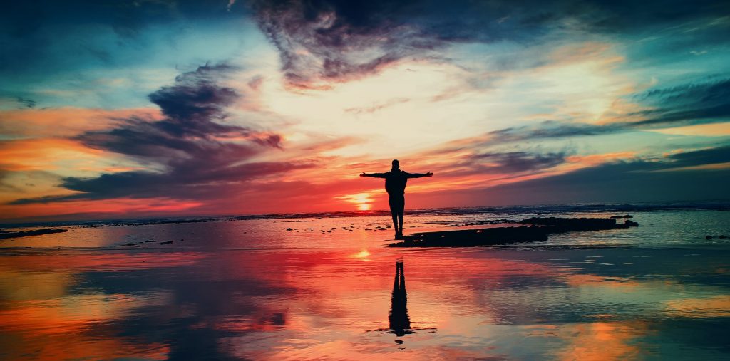 Photo of beautiful sunset on the water with man in foreground standing with arms outstretched.
