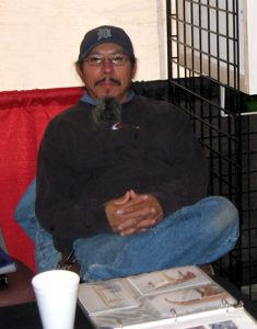 First Nations middle aged man sporting a moustache and longer goatee. He is sitting in front of a coffee table that has a styrofoam cup and an opened photo album sitting on it. His one leg is crossed, hands clasped in his lap. He is wearing glasses, a baseball cap,a black sweater and blue jeans.