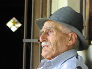 Profile of an elderly man seated against a window wearing a gray fedora hat and a light blue dress shirt.