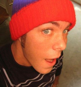 Close-up of a young man with grey eyes and a surprised expression wearing a red and blue toque and a white striped black sweater.