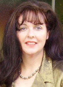 A photograph of a smiling woman mid-thirties, fair complexion, with long chestnut coloured hair with bangs, sporting a burnished gold jacket and a gold-beaded necklace.