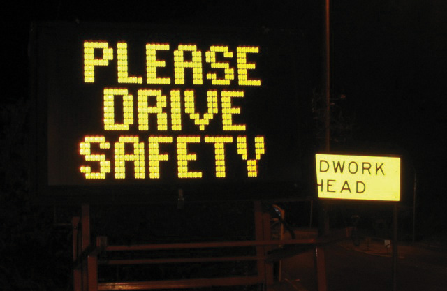 A lit up road sign saying please drive safety dworkhead