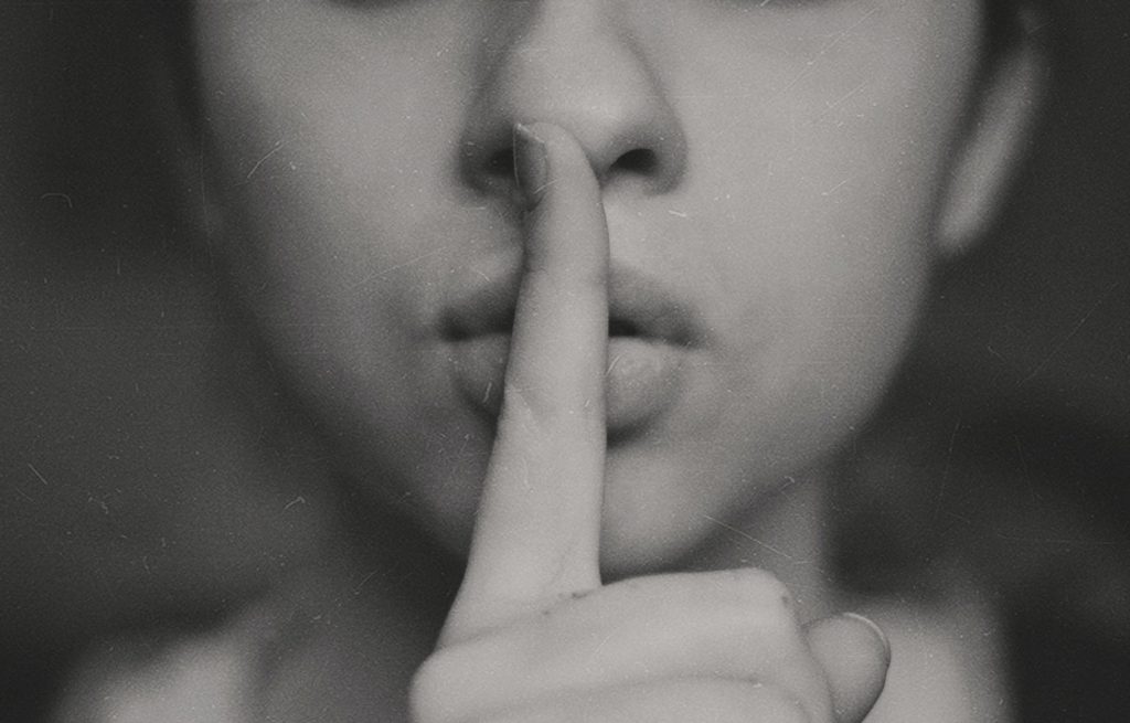 A person raises their index finger to pursed lips, indicating a "quiet" or "shh" gesture.