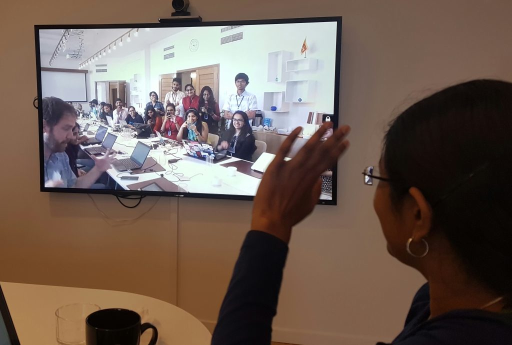 A person waves at a webcam attached to a video monitor, interacting with a classroom of students.