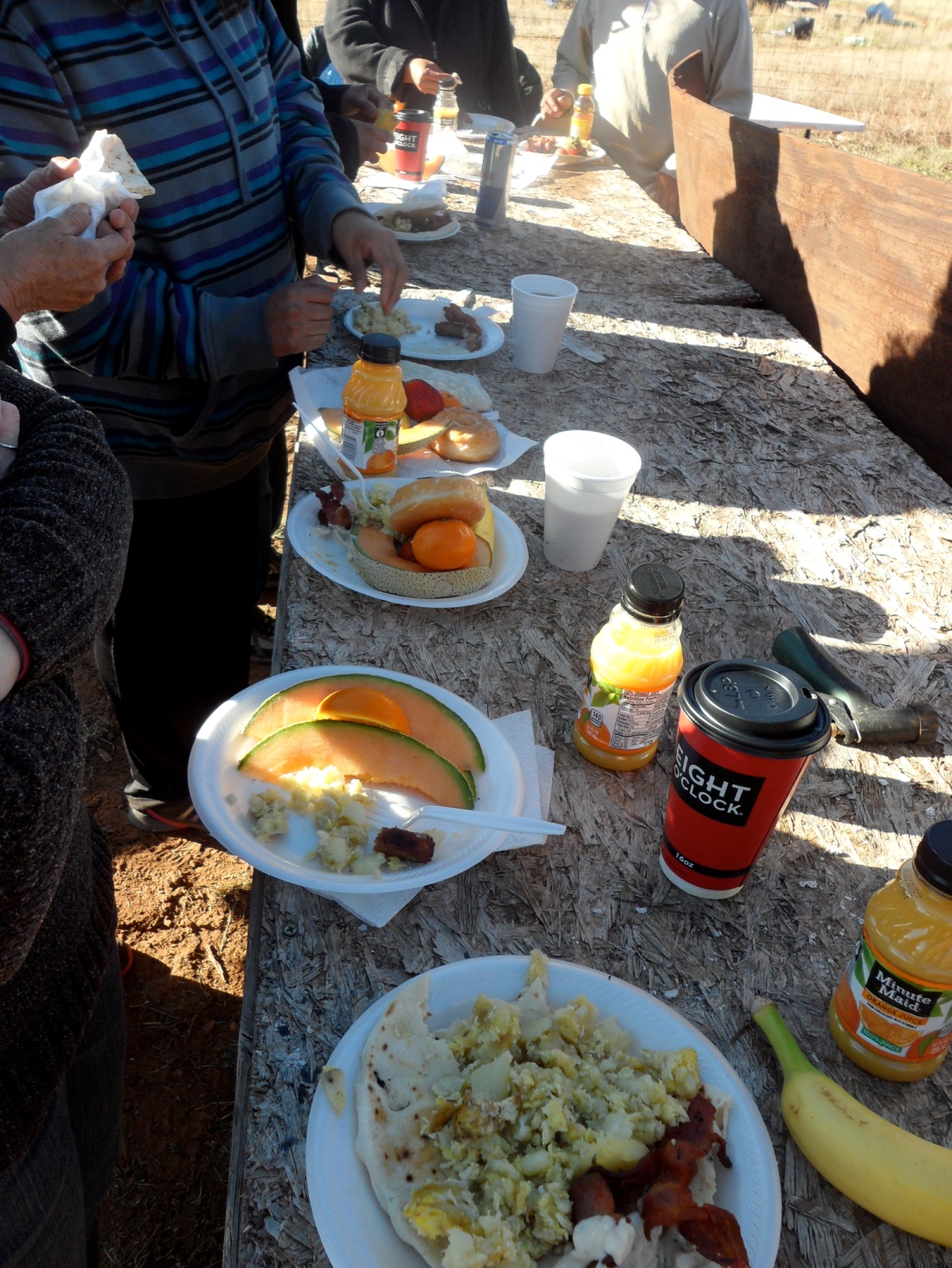 people standing around an outdoor table with many plate of food on it