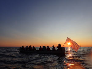 People in a canoe on open water at sunset with a flag reading "Délįnę Got'įnę Government"