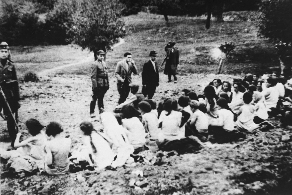 German police and auxiliaries in civilian clothes look on as a group of undressed Jewish women sitting on the ground before their execution.