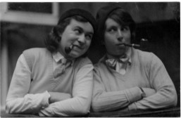Annette Eick (right) and her partner, Gertrude Klingel (left) pose in matching outfits; each wears a knitted vest over a collared shirt, a beret, a tie and sports a tobacco pipe hanging from their mouths.  