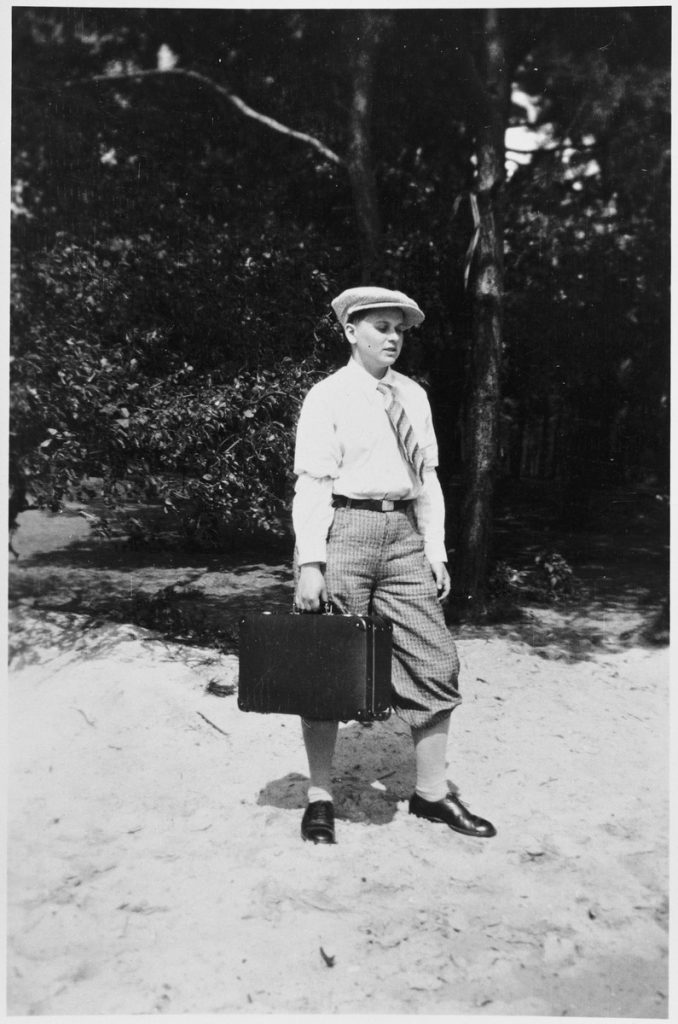 Gerd Katter stands in the sand against a backdrop of trees, looking down and to the right. He is wearing a collared shirt, slacks, shiny dress shoes, a striped tie and a cap. He carries a briefcase.  