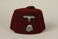 From the physical description provided by the United States Holocaust Museum Memorial: Red felt fez with a black tassel. The front of the cap has two patches embroidered in silver thread: the top patch is in the shape of a Reichsadler, a sinistra eagle with outstretched wings holding a wreath encircling a swastika in its talons. Below this is a Totenkopf [a Death’s head], or skull and crossbones.