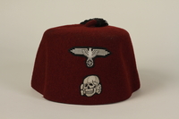 From the physical description provided by the United States Holocaust Museum Memorial: Red felt fez with a black tassel. The front of the cap has two patches embroidered in silver thread: the top patch is in the shape of a Reichsadler, a sinistra eagle with outstretched wings holding a wreath encircling a swastika in its talons. Below this is a Totenkopf [a Death’s head], or skull and crossbones. 