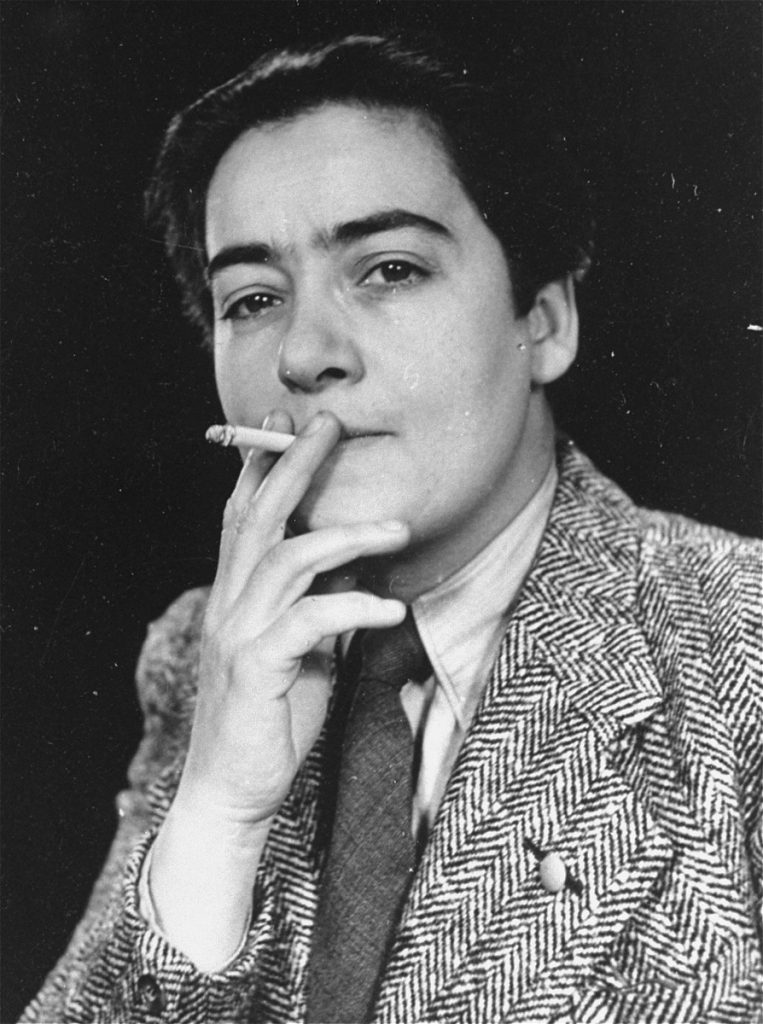 Frieda Belinfante smoking a cigarette in a blazer and tie, her hair is slicked back. 