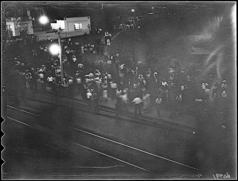 A black and white image of a crowd of people gathered on a street at night.