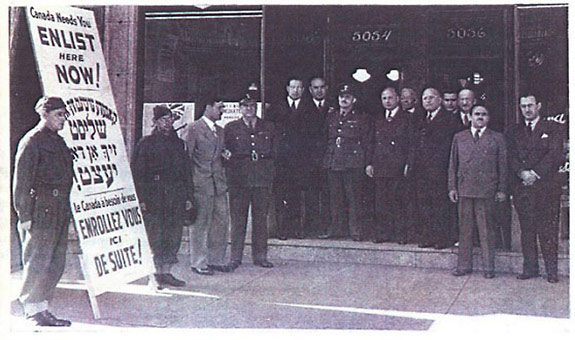 A group of men dressed in military uniforms and suits stand in front of a building with a sign that is written in English, Yiddish and French to attract potential army recruits.