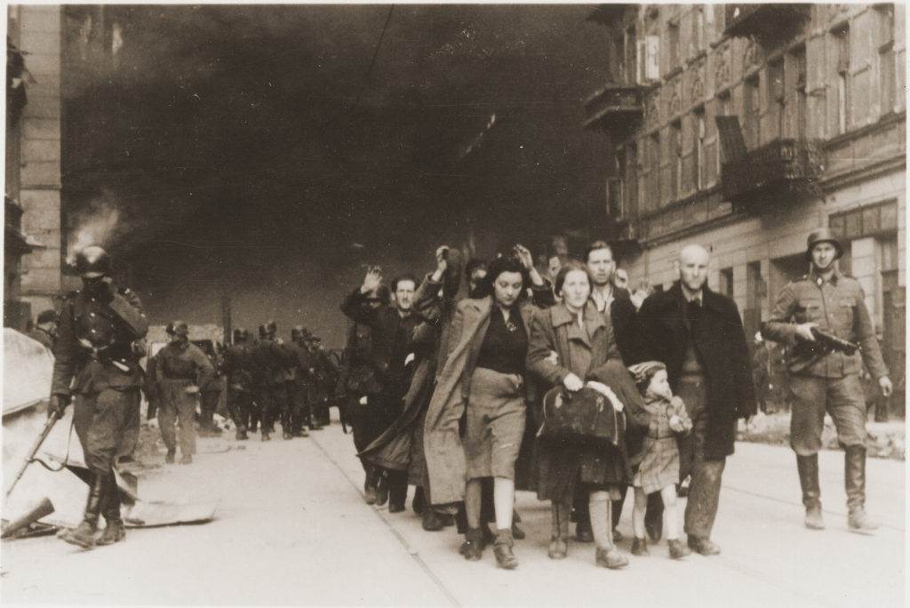 Jews captured during the suppression of the Warsaw ghetto uprising are led away from the burning ghetto by SS guards. The original caption (translated from German) reads, "Pulled from the bunkers by force." -United States Holocaust Memorial Museum