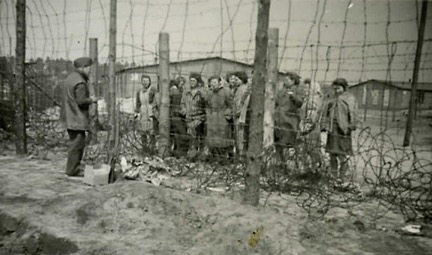 A man talks to a group of prisoners separated by two wire fences.