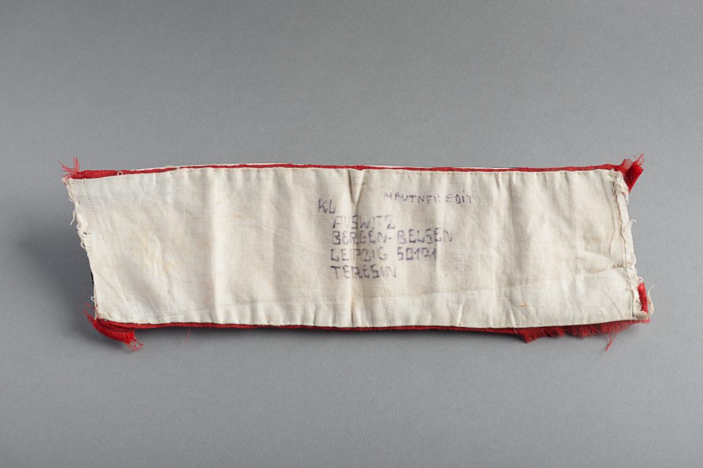 A red and white armband inscribed on the interior with the names of four concentration camps: Auschwitz; Bergen-Belsen; Leipzig; and Terezin. —Montreal Holocaust Museum.