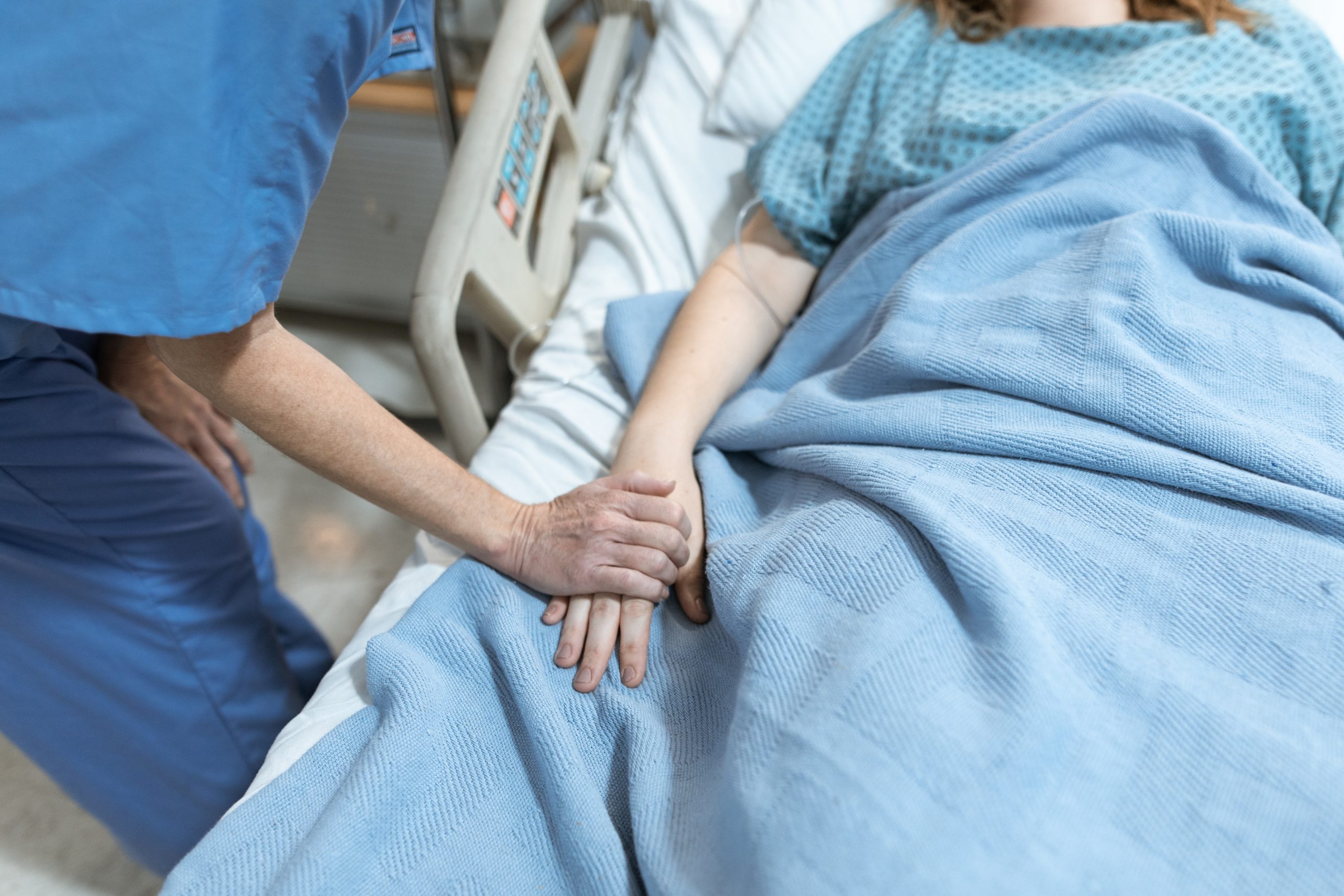 woman in scrub suit leans holds hand of a young patient lying in a hospital bed. The patient is wearing a gown and has a blanket.