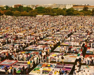 A grass field filled with many quilts and people standing between them to create an enormous patchwork quilt memorial for people who died of AIDs-related causes.
