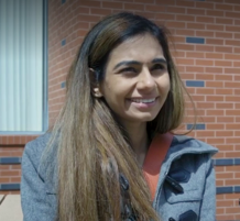 South Asian woman with long brown hair. She is smiling and wearing a blue duffle coat and peach coloured shirt.