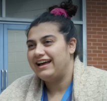 Biracial individual with long black hair pulled into a bun on top of their head. They are smiling and wearing a beige cardigan over blue hospital scrubs.