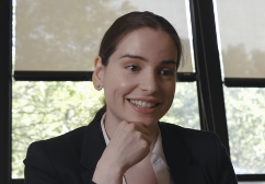 White woman with long brown hair pulled back in a low poney tale. She is smiling with her chin resting in her hand and wearing a black suit jacket over a white collared shirt.