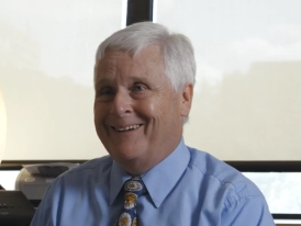 White male with short white hair. He is smiling and wearing a blue collared shirt and tie.