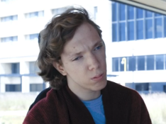White, non-binary person with short blonde wavey hair. They are looking serious and wearing a burgundy poncho and blue t-shirt.