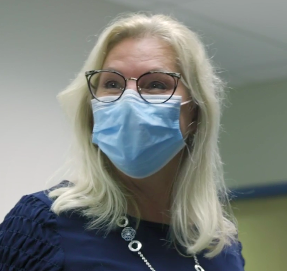 White woman with white/blonde should-length hair and glasses. She is wearing a mask and a blue top with a long silver necklace.
