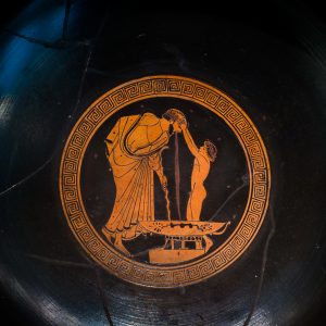 Red-Figure Attic kylix from approximately 490 BCE depicting an upper-class man throwing up into a bowl after drinking too much at a symposium. A young, nude slave boy is holding the man's head/hair and assisting him while he is throwing up.