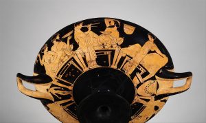 Terracotta Red-Figure Kylix from Athens around 480 BCE. The drinking cup depicts symposia imagery: from left to right a woman and man sitting together nearly nude. The woman is likely a slave or hetaira based on her presence at a symposium. The man with her is holding a kylix and is drinking wine out of it. The middle of the vessel depicts a young boy, likely a slave, playing the flute. He attended the symposium in order to entertain the other guests. Behind the slave is a man reclined on a klinai - the benches that were used at symposia. He looks relaxed and as though he is enjoying the entertainment the young boy is providing. He is holding a drinking vessel and drinking wine. To the far right there is a woman. Some of the image does not survive, but it looks like she is massaging a man sitting on a klinai. Surrounding the main images are various objects, including amphoras, kylixes, and various other vessels.